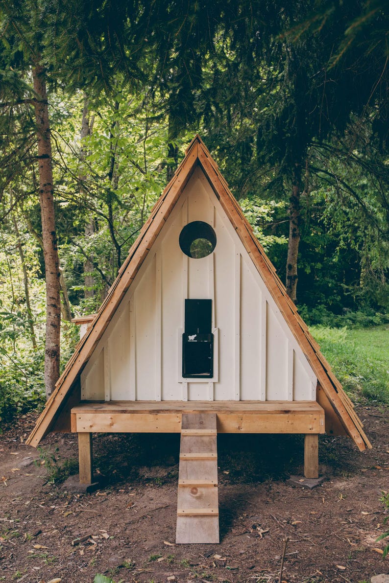 May include: A-frame chicken coop with a white exterior and a wooden roof and trim. The coop has a round window, a black door, and a small wooden ramp leading to a platform. The coop is set in a wooded area.