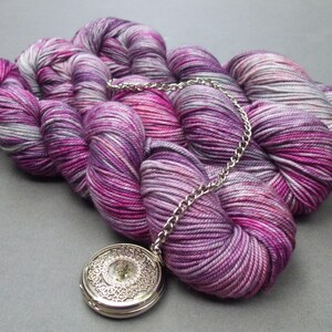 May include: A skein of hand-dyed yarn in shades of purple, pink, and grey, with a silver chain and a decorative silver locket. The yarn is arranged in a loose, flowing shape, and the locket is positioned in the foreground.