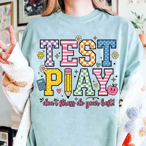 Puede incluir: Camiseta azul claro con las palabras "TEST PLAY" en letras coloridas y estampadas. El dise&ntilde;o incluye un l&aacute;piz, una calculadora, una manzana y la frase "don't stress do your best!" en una fuente l&uacute;dica. La camiseta tiene un corte holgado.