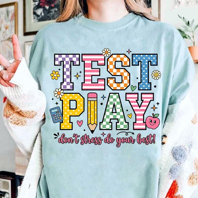 Puede incluir: Camiseta azul claro con las palabras "TEST PLAY" en letras coloridas y estampadas. El dise&ntilde;o incluye un l&aacute;piz, una calculadora, una manzana y la frase "don't stress do your best!" en una fuente l&uacute;dica. La camiseta tiene un corte holgado.