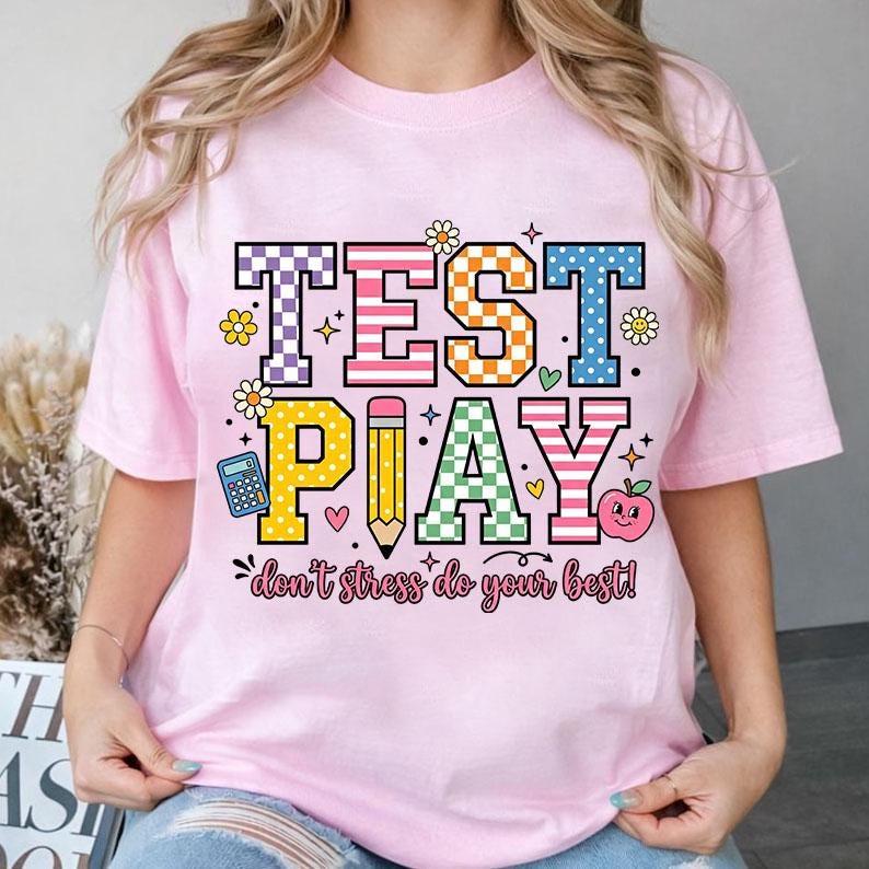 Puede incluir: Camiseta rosa claro con las palabras "TEST PLAY" en letras coloridas y estampadas. El dise&ntilde;o incluye una calculadora, un l&aacute;piz, una manzana y la frase "don't stress do your best!"