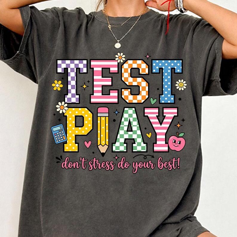 Puede incluir: Camiseta gris oscuro con un gr&aacute;fico colorido que dice "TEST PLAY" en letras grandes. El dise&ntilde;o incluye un l&aacute;piz, una manzana, una calculadora y la frase "don't stress do your best!"