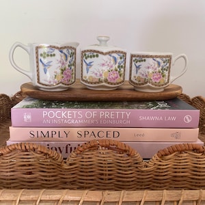 May include: A white ceramic creamer, sugar bowl with lid, and teacup set on a wooden tray. The set features a floral and bird design with pink, blue, and green accents. The set is displayed on top of a stack of books.