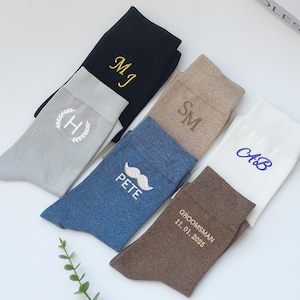 Può includere: Un assortimento di calzini personalizzati in vari colori, tra cui blu navy, grigio, beige, blu e bianco. Ogni paio presenta ricami personalizzati con iniziali, una grafica di baffi o la parola "Groomsman" con una data. I calzini sono disposti su una superficie bianca.