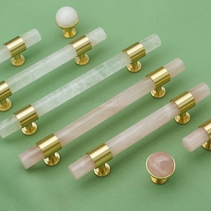 May include: A collection of cabinet hardware, including crystal and rose quartz drawer pulls and knobs. The pulls have a cylindrical design with gold-toned metal accents. The knobs are round with a gold base. The hardware is arranged on a green surface.