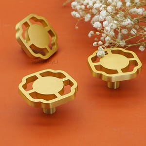 以下が含まれることがあります： ユニークな透かし彫りデザインの、3つの金色のキャビネットノブ。各ノブは、装飾的なスカラップフレームに囲まれた中央の円形要素を備えています。ノブはオレンジ色の背景に表示され、背景には白い花があります。