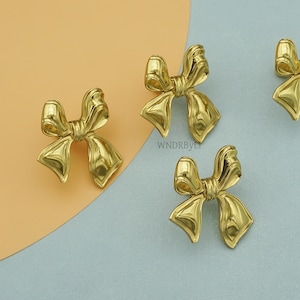 May include: Four gold-colored bow-shaped decorative objects. The bows are highly reflective, with a smooth, polished surface. The background is a split of light orange and light blue. The text "WNDRBYLT" is visible on the background.