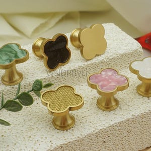 May include: Six decorative cabinet knobs with a cloverleaf design. The knobs have gold-colored bases and tops in various colors and patterns, including green, black, gold, pink, and white. The knobs are displayed on a textured surface.