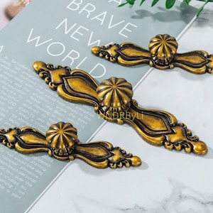 Può includere: Tre maniglie per mobili antiche color oro con disegni floreali ornati. Ogni maniglia presenta un pomello centrale rotondo ed elementi terminali decorativi. Le maniglie sono disposte su una superficie grigio chiaro con testo sullo sfondo.