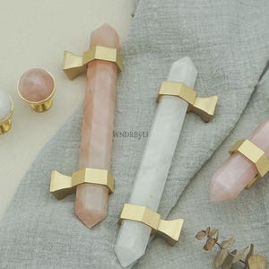 Peut inclure: Ensemble de boutons et poignées de meubles en cristal. Les boutons sont ronds avec des bases dorées, tandis que les poignées sont des cristaux allongés roses et blancs, maintenus par des supports en métal doré. Sur un tissu gris.