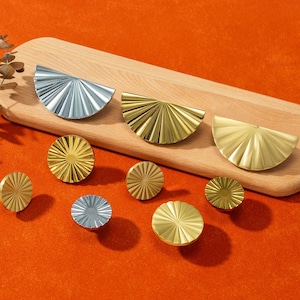 Może przedstawiać: Kolekcja dekoracyjnych okuć do szafek w odcieniach złota i srebra. Okucia obejmują wzory półkoliste i okrągłe z radialnym wzorem, prezentowane na drewnianej desce na pomarańczowym tle.