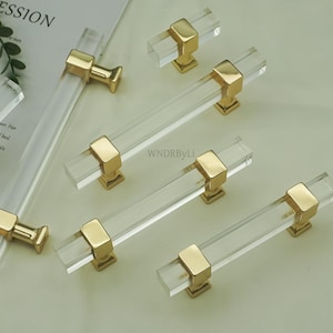 May include: Clear acrylic cabinet pulls with gold-toned hardware. The pulls are rectangular and have a modern design. The hardware is square and connects the acrylic bar to the cabinet. The pulls are arranged on a light-colored surface.