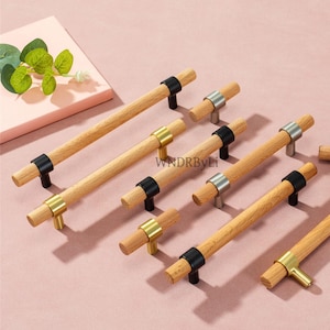 May include: A collection of wooden cabinet pulls with various metal accents. The pulls feature natural wood handles with black, gold, and silver-toned metal details. The hardware is arranged on a pink surface.