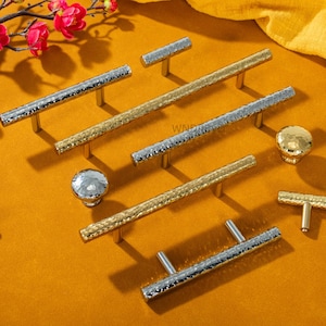 May include: An assortment of silver and gold cabinet hardware, including handles and knobs, with a hammered texture. The hardware is displayed on a mustard-colored surface, with a sprig of pink flowers in the upper left corner.