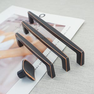 Op de afbeelding: Een set bronzen kastbeslag, waaronder drie rechthoekige stanggrepen en een ovale knop. De grepen hebben een donkere afwerking met koperkleurige randen. De hardware wordt gepresenteerd op een tijdschriftpagina.