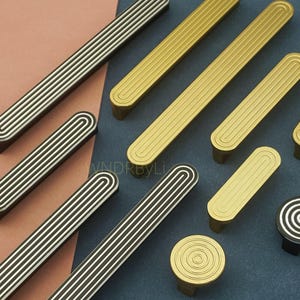 May include: An assortment of cabinet hardware in various styles and finishes. The image features long, rectangular pulls with grooved detailing in black and silver, and gold. Round knobs with concentric circles are also visible.