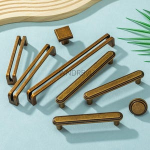 May include: A selection of antique brass cabinet handles and a square knob, displayed on a light blue surface. The collection includes different handle designs and a round knob. The handles vary in length, with the longest being approximately 30 cm.
