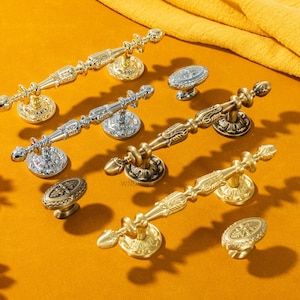 Puede incluir: Una variedad de tiradores y pomos decorativos para armarios, con acabados dorados y plateados. La ferretería presenta intrincados diseños florales y de volutas. Los artículos se muestran sobre una superficie amarilla. Medidas en centímetros.