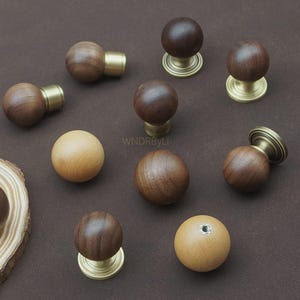 Könnte beinhalten: Eine Auswahl an Holzmöbelknöpfen in verschiedenen Ausführungen. Einige sind kugelförmig mit Messingbasen, andere zylindrisch mit Messingakzenten. Die Holztöne reichen von hell- bis dunkelbraun und zeigen natürliche Maserungsmuster. Die Knöpfe sind auf einer braunen Oberfläche angeordnet.