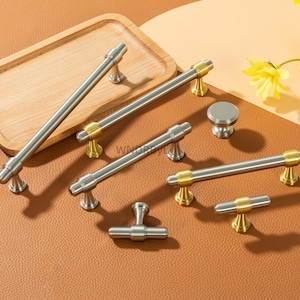 May include: A collection of silver and gold cabinet hardware, including handles and a knob, arranged on a brown surface. The hardware features a brushed silver finish with gold accents. A wooden tray and yellow flowers are in the background.