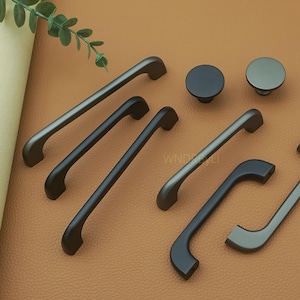 May include: A selection of matte black cabinet hardware, featuring various pull handles and two round knobs. The handles come in different shapes and sizes, with both curved and straight designs. The hardware is presented on a brown surface.