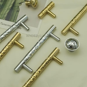 May include: An assortment of cabinet hardware, including silver and gold-toned T-bar handles and a faceted knob. The handles have a textured, hammered appearance. The image also shows a small gold knob. The text "Tenacious life" is visible in the background.