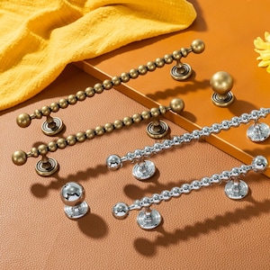 May include: A selection of cabinet hardware, including bronze and silver-toned pulls and knobs. The pulls have a beaded design, while the knobs are spherical. The hardware is displayed on a brown surface with a yellow cloth and a yellow flower.