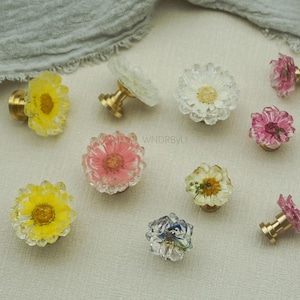 Puede incluir: Un conjunto de tiradores decorativos para armarios con flores prensadas reales encapsuladas en resina transparente. Los tiradores tienen herrajes de latón y están disponibles en varios colores, como amarillo, rosa, blanco y azul.