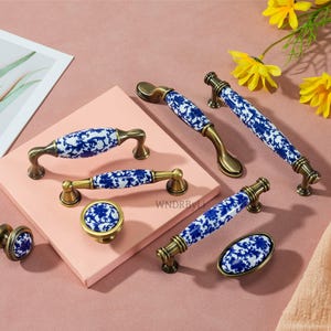 Könnte beinhalten: Eine Sammlung dekorativer Möbelbeschläge mit blau-weißem Blumendekor auf Keramikgriffen. Die Beschläge umfassen Knöpfe, Griffe und Ziehgriffe mit antiken messingfarbenen Metallbasen. Die Artikel sind auf einer rosa Oberfläche angeordnet.