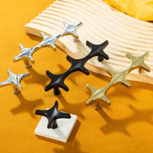 May include: A collection of star-shaped cabinet pulls in silver, black, and gold finishes. The hardware is displayed on a wooden surface and a square stone tile against a yellow background. The pulls have a modern design.