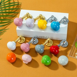 May include: A collection of colorful cabinet knobs in various shades, including pink, yellow, blue, and green. The knobs have a rounded shape and are attached to gold or silver hardware. The knobs are displayed on a white surface against a warm orange background.