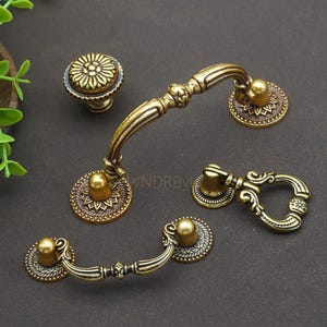 Puede incluir: Un surtido de herrajes de gabinete de latón antiguo, que incluye un pomo, dos tiradores y un tirador decorativo. Los herrajes presentan intrincados diseños florales y de volutas, con un acabado cálido y envejecido. Las piezas están dispuestas sobre una superficie oscura.