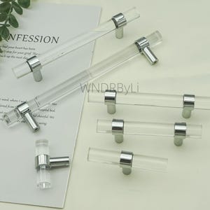 May include: Clear acrylic cabinet pulls with polished chrome hardware. The pulls are cylindrical with a T-bar design. Various sizes are shown, including a small knob and longer pulls. The background includes text from a magazine.