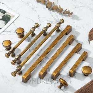 Può includere: Un assortimento di ferramenta per mobili in ottone antico, tra cui pomelli e maniglie. L'hardware presenta una finitura calda e invecchiata e vari design, dalle semplici maniglie a barra a pomelli e maniglie ornate e dettagliate. L'hardware è disposto su una superficie di marmo.