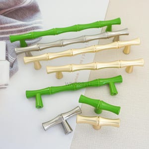 以下が含まれることがあります： さまざまな色の竹スタイルのキャビネットプル。プルは緑、シルバー、ゴールドで、T字型のハンドルが付いています。竹のデザインは、家具に自然でエレガントなタッチを加えます。引き出しやキャビネットに最適です。