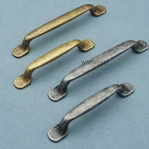 May include: Four antique-style cabinet pulls in bronze and silver finishes. The pulls have a curved handle design with decorative end pieces. The image shows the pulls against a blue background.