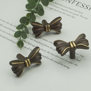 May include: Three bronze-coloured bow-shaped cabinet knobs with gold accents. The knobs are arranged on a white surface with text and a green plant. These decorative knobs add a touch of elegance to furniture.