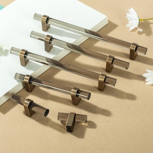 Może przedstawiać: Kolekcja przezroczystych akrylowych uchwytów do szafek z podstawami z metalu w kolorze brązu. Uchwyty są cylindryczne i prostokątne, o nowoczesnym designie. Okucia są ułożone na neutralnej powierzchni, prezentując różne rozmiary i style.