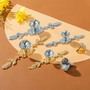 Op de afbeelding: Decoratieve kastbeslagen met kristallen knoppen en sierlijke metalen achterplaten. De hardware is verkrijgbaar in zilveren en gouden afwerkingen. De kristallen knoppen voegen een vleugje elegantie toe aan elke kast of lade.