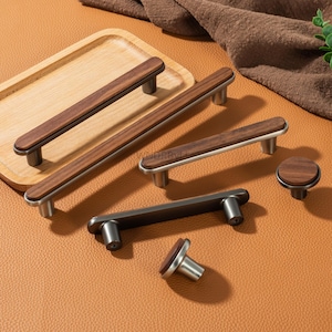 May include: A collection of cabinet hardware, including handles and knobs. The handles feature a dark wood center with silver or dark gray metal accents. The knobs have a matching wood and metal design. The hardware is displayed on a brown surface.