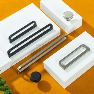 Może przedstawiać: Asortyment okuć do szafek, w tym czarne i srebrne uchwyty, srebrny gałka i czarny gałka. Uchwyty mają nowoczesny, minimalistyczny design. Okucia są prezentowane na białych blokach na żółtym tle.