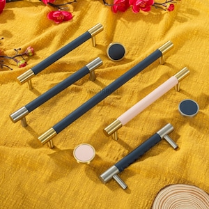 Può includere: Set di ferramenta per mobili con maniglie e pomelli blu navy, rosa e argento, con dettagli dorati e argentati. Le maniglie hanno una consistenza simile alla pelle. L'hardware è esposto su una superficie gialla.