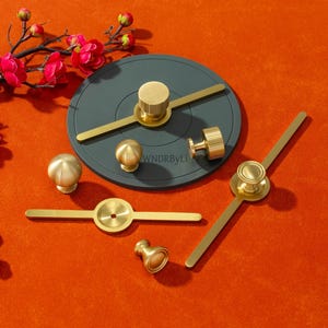 Op de afbeelding: Een verzameling messing kastbeslag, waaronder knoppen en handgrepen, gepresenteerd op een oranje oppervlak. De hardware omvat een cirkelvormig accentstuk. Rode bloemen op de achtergrond. Afmetingen in cm.