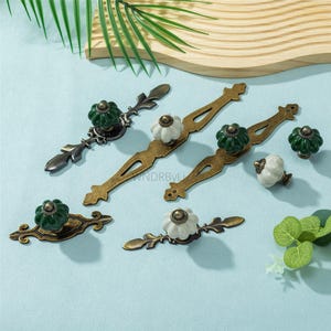 May include: A collection of decorative cabinet hardware, including drawer pulls and knobs. The pulls have ornate, antique-style designs with brass-colored bases and green or white ceramic knobs. The hardware is arranged on a light blue surface.