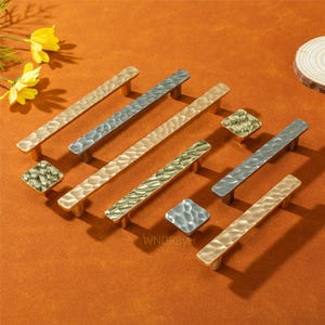 Pode incluir: Uma variedade de puxadores e puxadores de armário em metal martelado em tons de ouro, prata e bronze. Os puxadores são retangulares com uma superfície texturizada, e os puxadores são quadrados. O fundo é laranja quente.