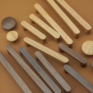 May include: A collection of wooden cabinet pulls and knobs in various shades of brown. The pulls are rectangular with rounded edges, and the knobs are circular. The wood grain is visible on the surface. The text "WNDRByLi" is visible.
