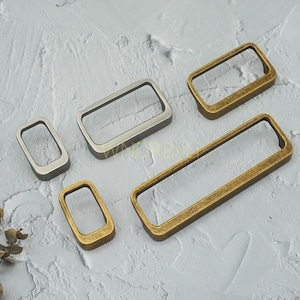 May include: An assortment of rectangular metal hardware in varying sizes and finishes. The hardware includes a brushed silver and several antique brass pieces. The pieces are open-framed, with a rectangular shape, and are likely intended for use as drawer pulls or cabinet handles.