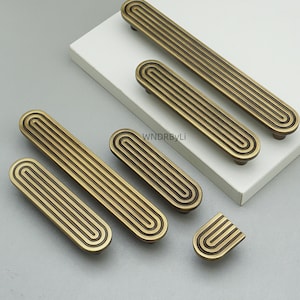 May include: A collection of antique brass cabinet pulls and knobs. The hardware features a grooved, oval design with rounded ends. The pulls vary in size, with a small knob included. The hardware is displayed on a white surface.