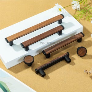 May include: A selection of wooden cabinet handles and knobs. The hardware has a dark wood grain finish with black detailing. The image includes rectangular pulls and round knobs. The text "WNDRByLi" is visible.