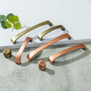 May include: A selection of cabinet handles and knobs in gold and copper finishes. The image displays curved handle pulls and round knobs in gold and copper tones, arranged on a white surface. The text "WNDRBYLT" is visible.